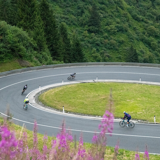 Arlberg Giro - Abfahrt vor Stuben Radfahrer fahren durch eine weite Kehre mit Bergwiesen und Wald – typische Arlberg-Giro-Szene